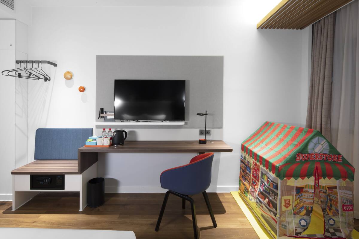 Ein modernes Hotelzimmer mit Fernseher, Schreibtisch mit Stuhl, einer kleinen Sitzecke und einem Spielhäuschen.