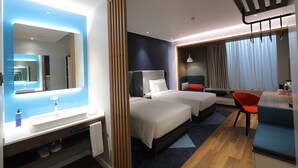 Une chambre d’hôtel avec un grand miroir, deux lits, un bureau avec une chaise, un lavabo et une fenêtre avec des rideaux.