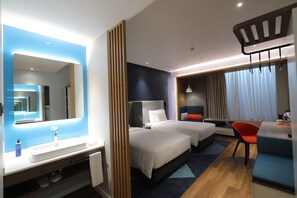 Hotelkamer met een grote spiegel, twee bedden, een bureau met stoel, een wastafel en een raam met gordijnen.