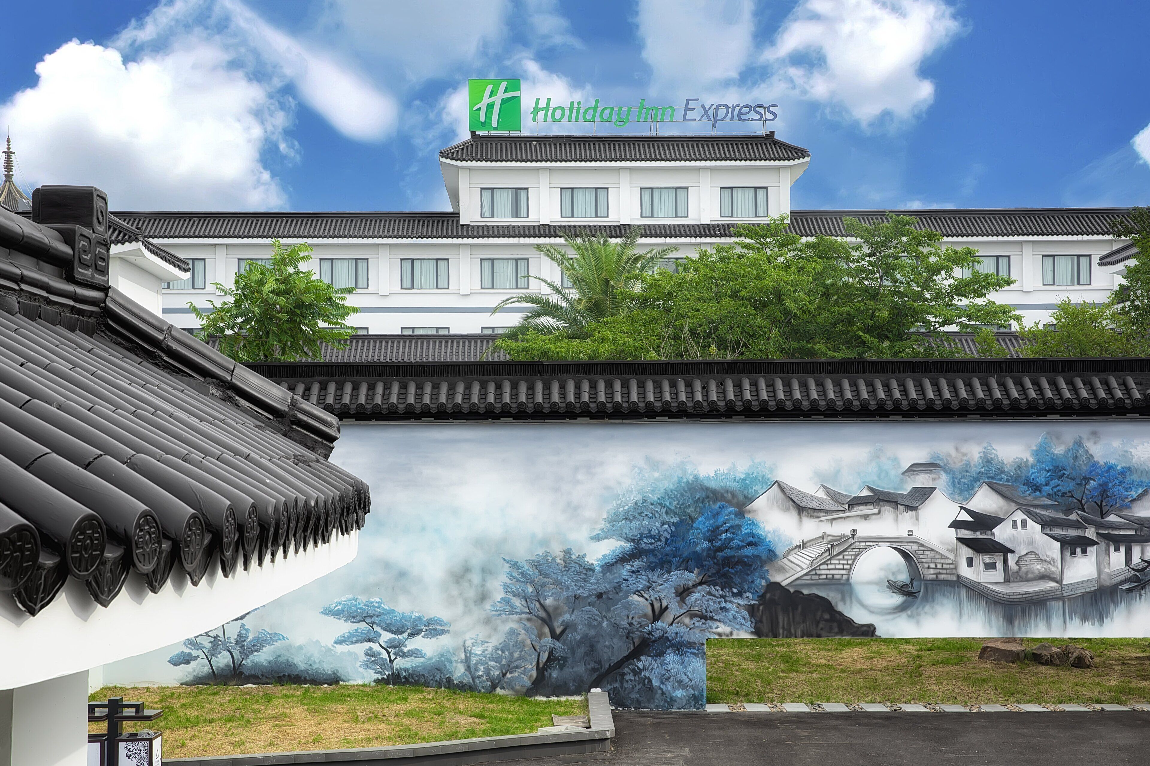 A traditional building with a modern Holiday Inn Express sign, a mural of a serene landscape, and a traditional tiled roof.