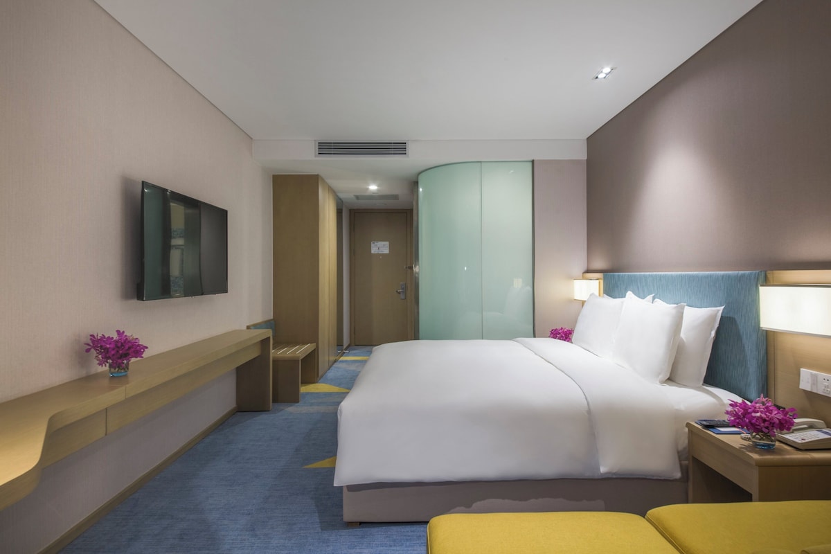 Quarto de hotel com cama, mesas de cabeceira, televisão, escrivaninha com luminária e porta do banheiro.