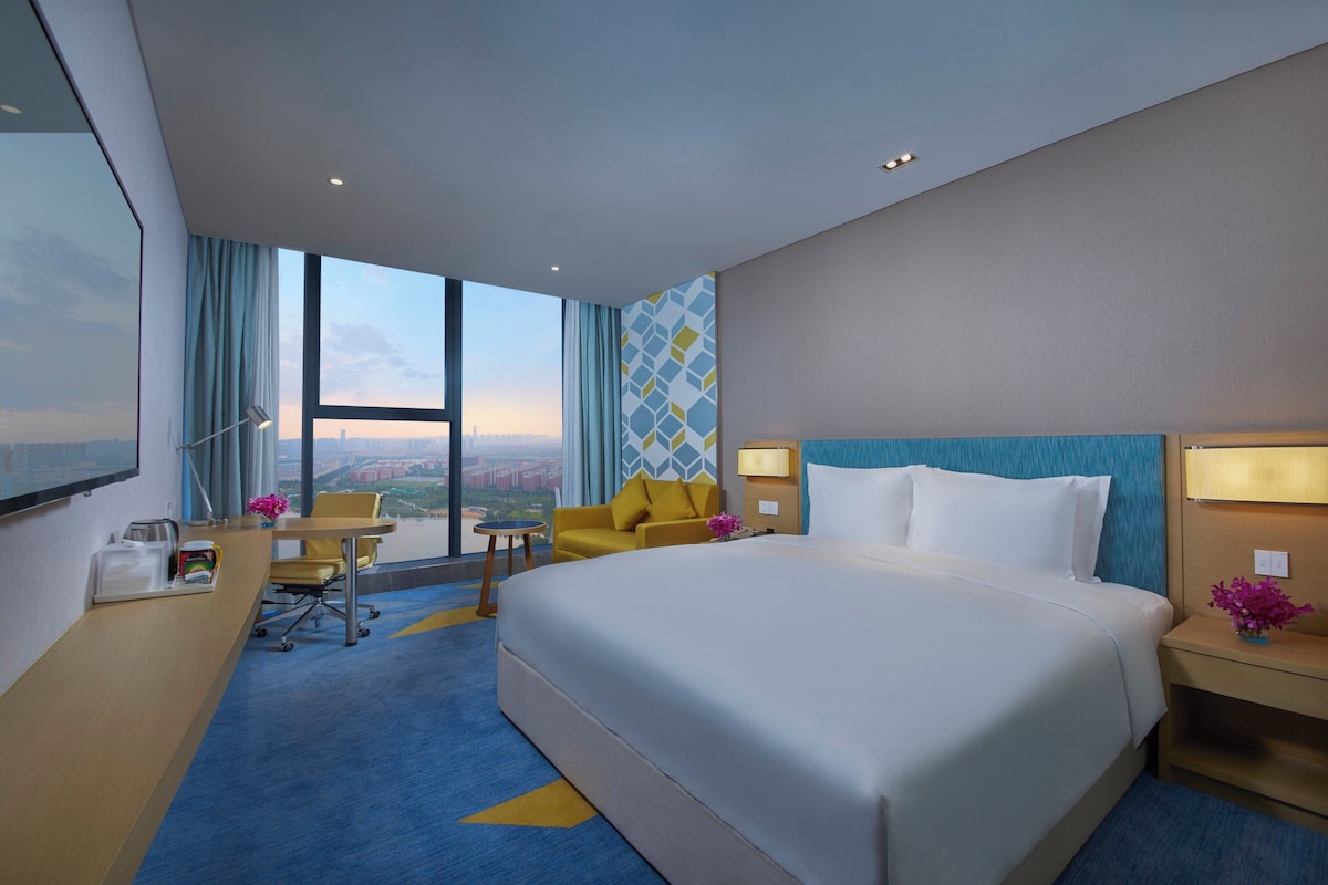 Quarto de hotel com uma cama grande, uma escrivaninha com cadeira, uma mesinha e vista para a cidade pela janela.