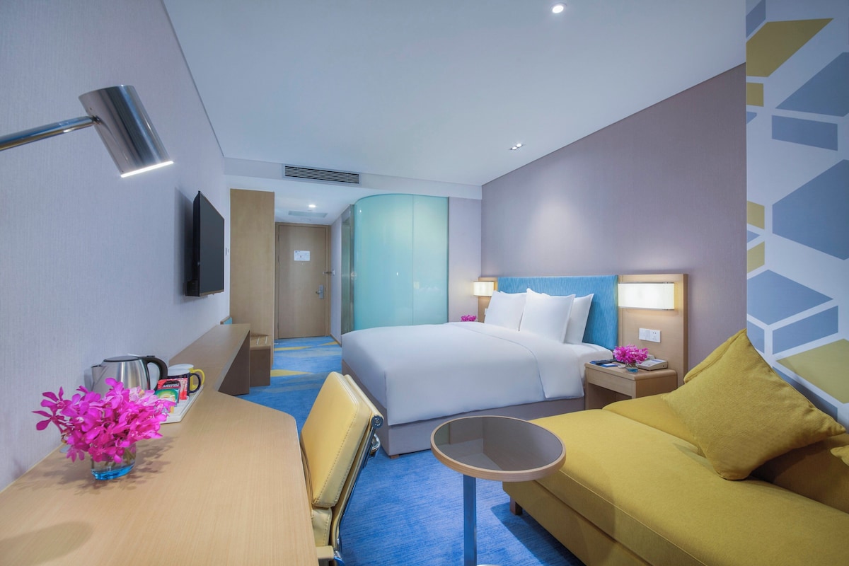 Quarto de hotel com cama, sofá, escrivaninha com luminária e televisão na parede.