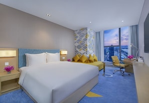 Quarto de hotel moderno com uma cama grande, uma escrivaninha, uma cadeira e vista para a cidade pela janela.