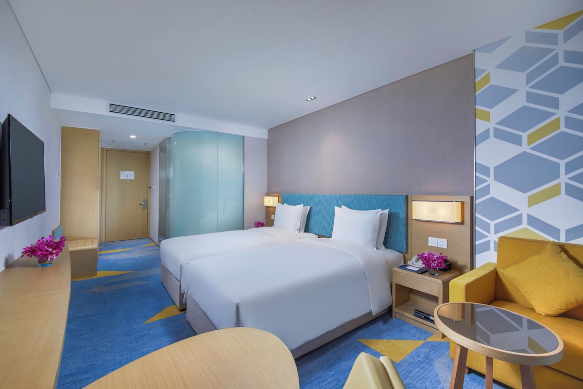 Quarto de hotel com cama, mesas de cabeceira, escrivaninha, cadeira e televisão.