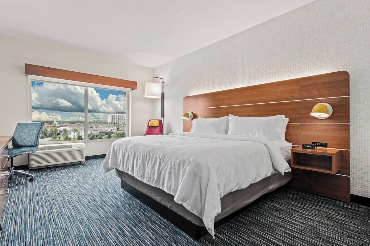 Ein Hotelzimmer mit einem großen Bett, einem Schreibtisch, einem Sessel, einem Fenster mit Blick auf die Stadt und einer Wandlampe.