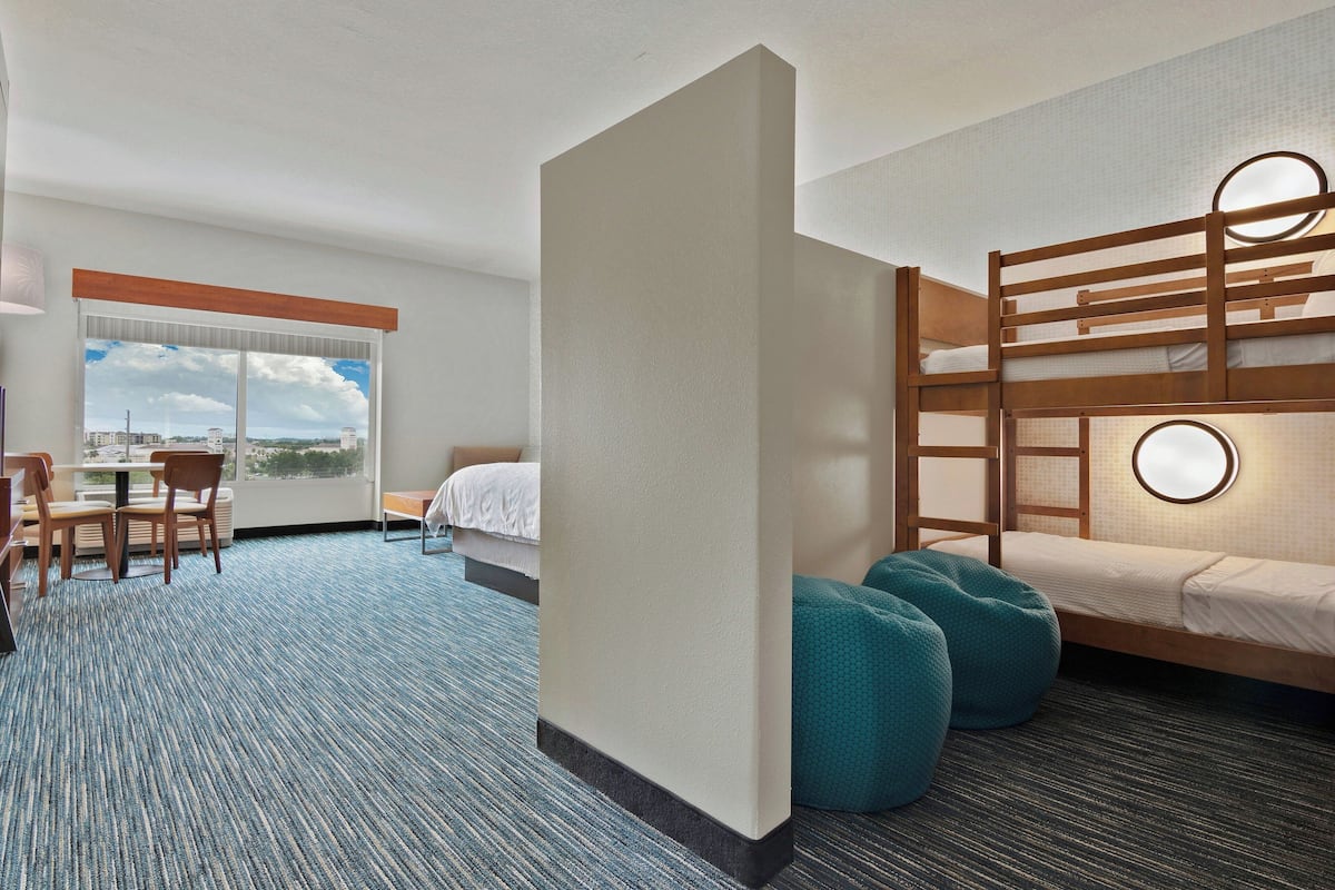 Ein Hotelzimmer mit einem Etagenbett, einem Schreibtisch mit Stühlen, einem großen Fenster mit Blick auf die Stadt und einem Teppichboden.