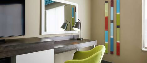 A hotel room with a desk, a chair, a flat-screen TV, a mirror, and a wall with colorful vertical stripes.