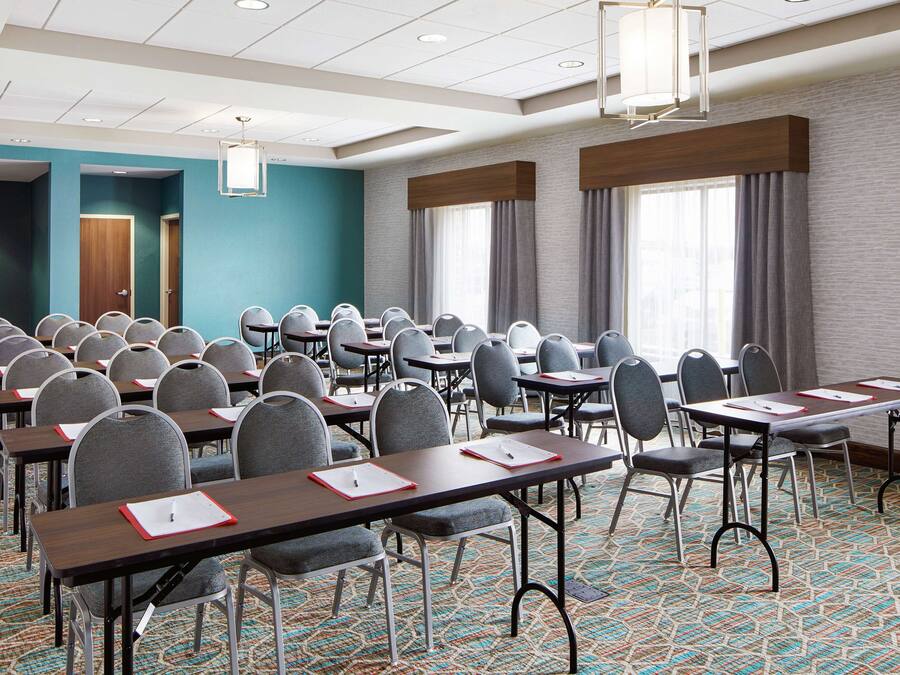 A conference room with rows of tables and chairs, large windows with curtains, and a patterned carpet.