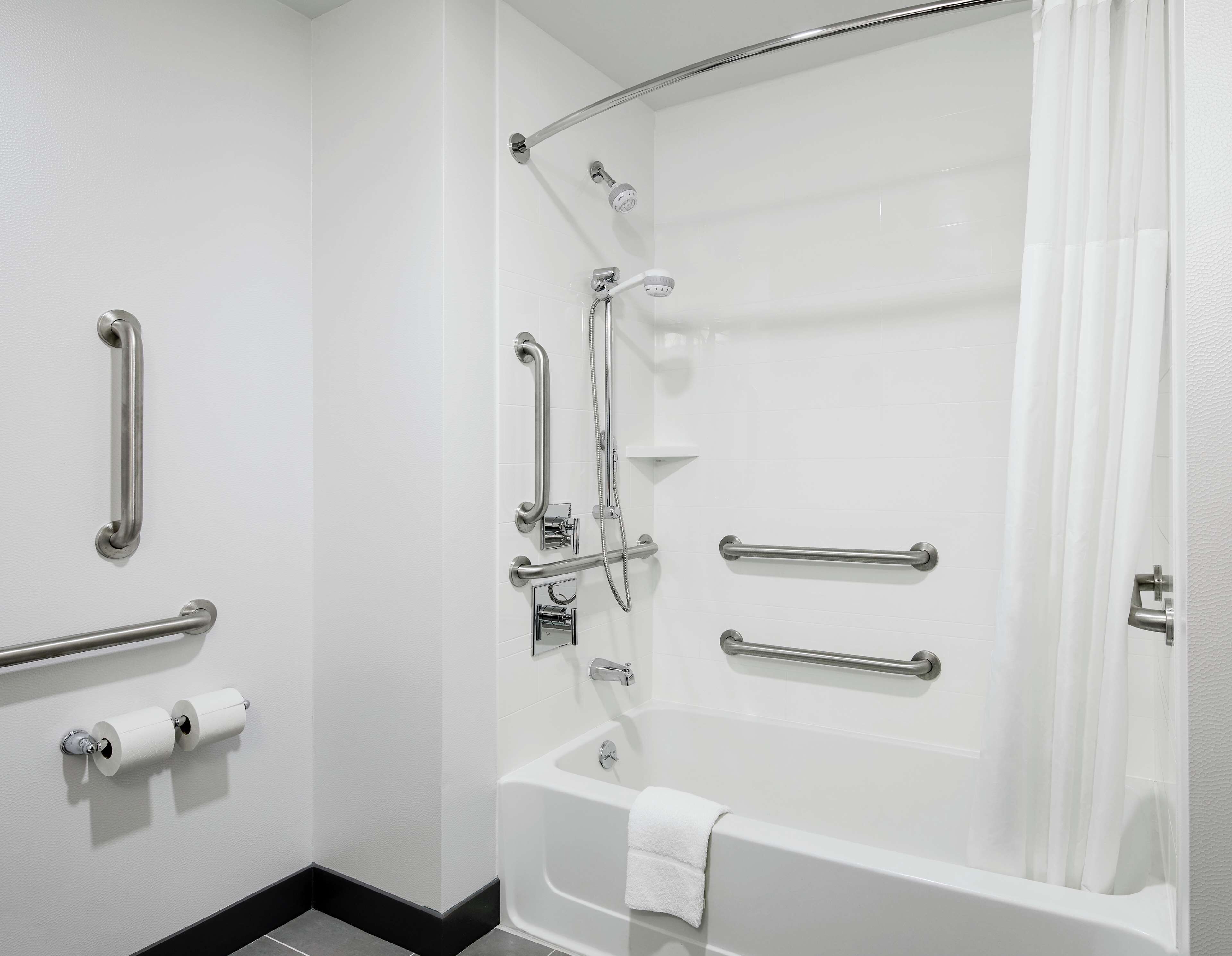 Un baño con bañera, ducha con barras de apoyo y soporte para papel higiénico.