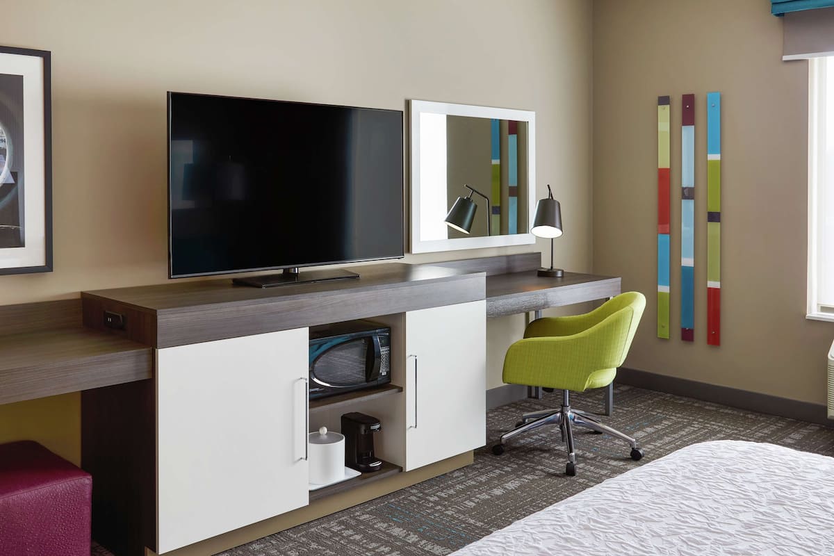A hotel room with a flat-screen TV, a desk with a lamp, a green chair, a coffee maker, and a bed.