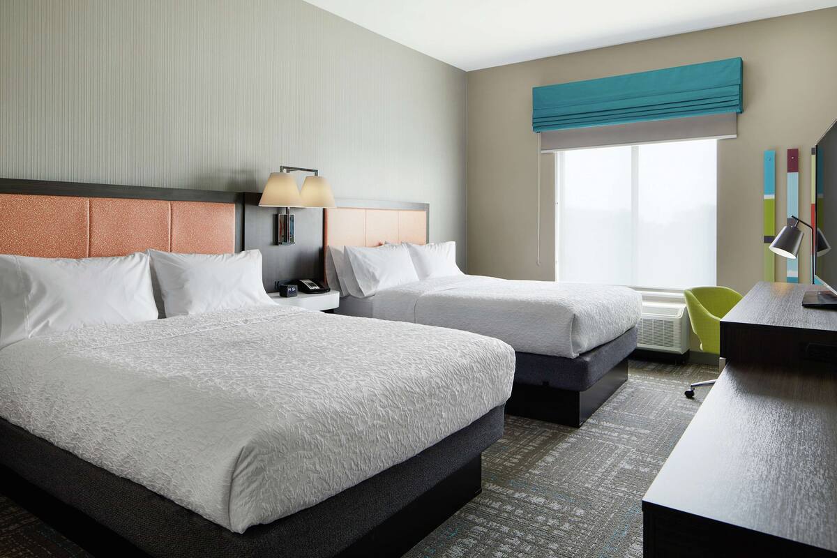 A hotel room with two beds, a desk, a chair, a lamp, and a window with a view.