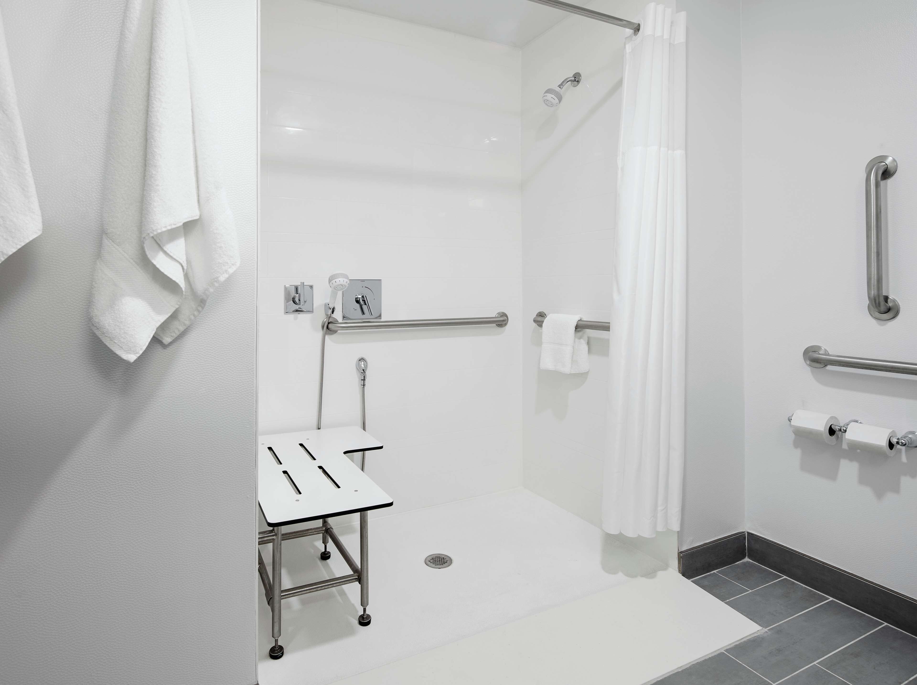 Un baño moderno con ducha, barras de apoyo, un dispensador de papel higiénico y un lavabo blanco con un perchero para toallas.