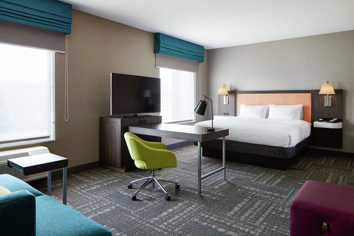 A hotel room with a bed, desk, chair, TV, lamps, and a window.
