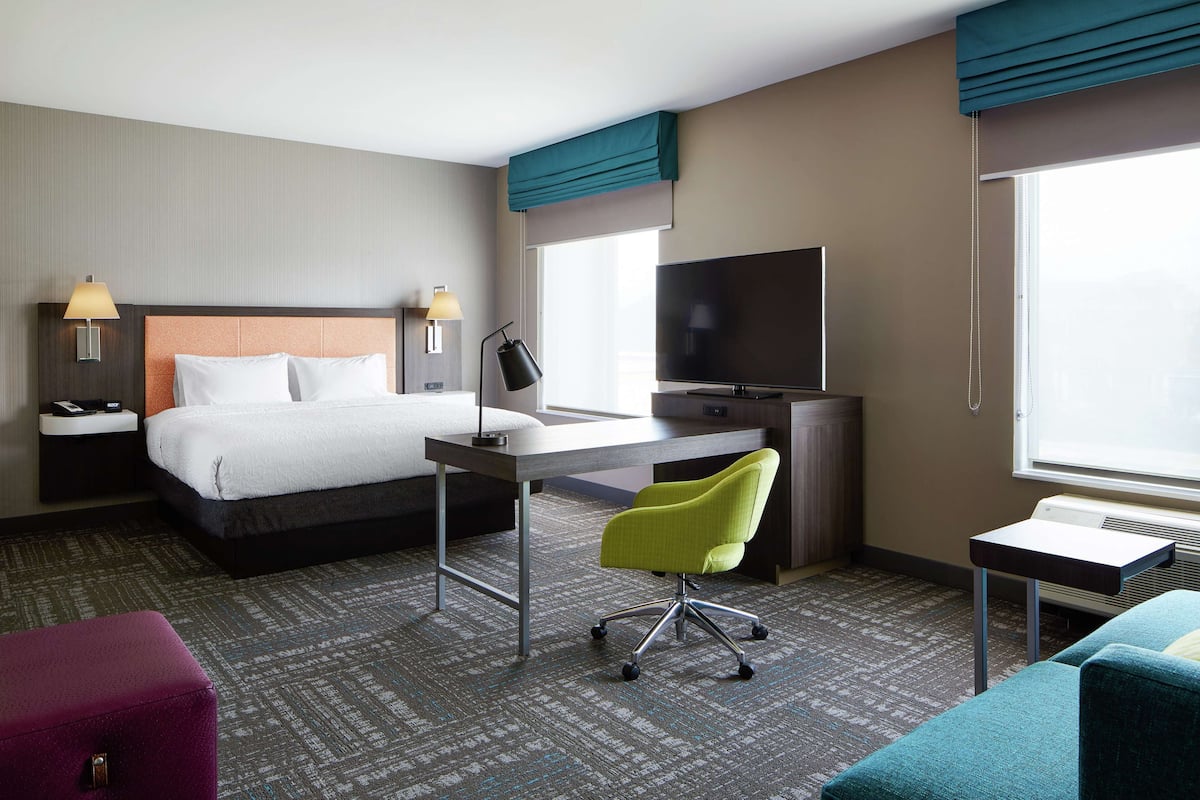 A hotel room with a bed, desk, chair, TV, and large windows.