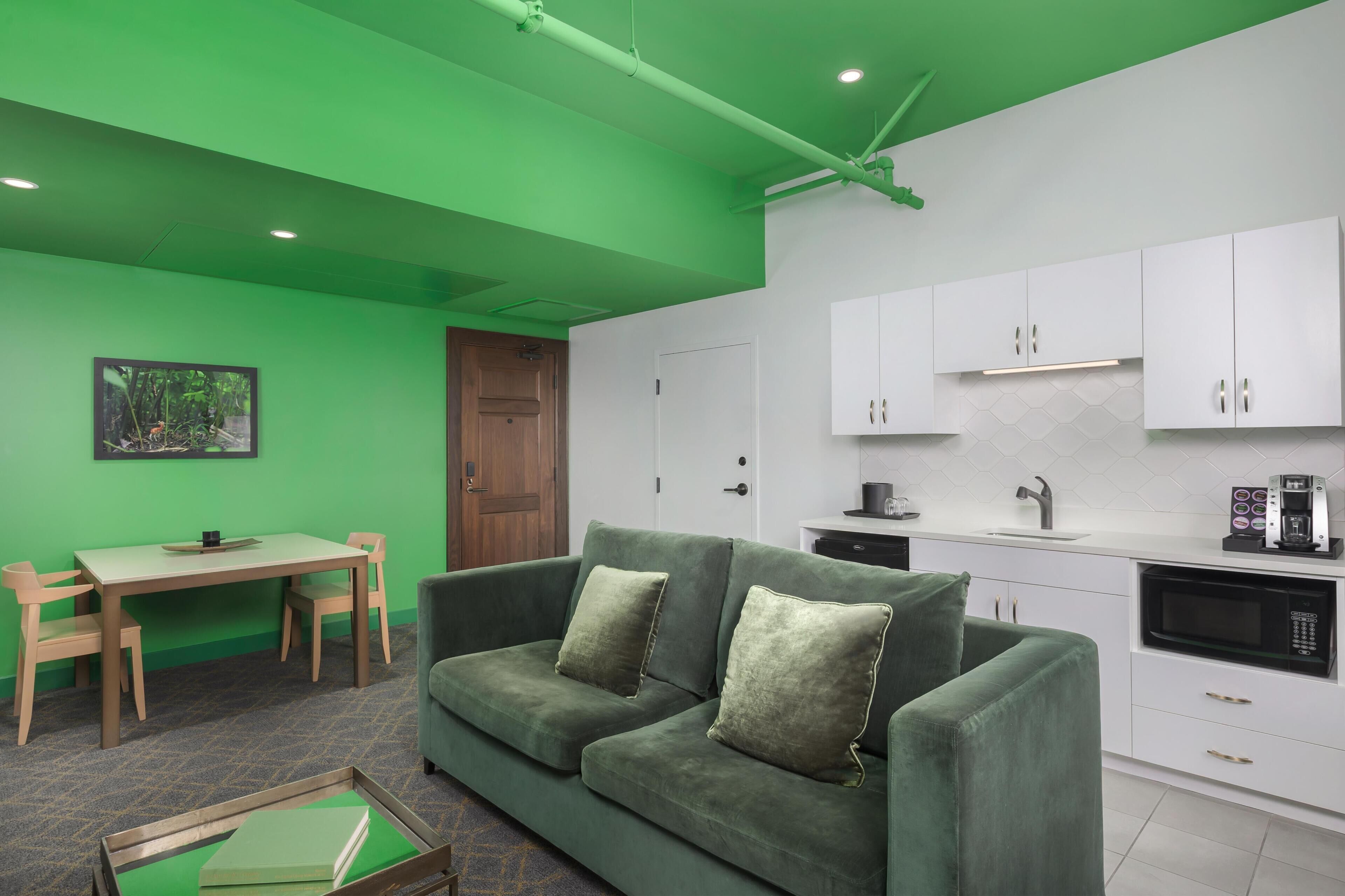 A modern living room with a green sofa, a dining table, and a kitchen area with white cabinets and appliances.
