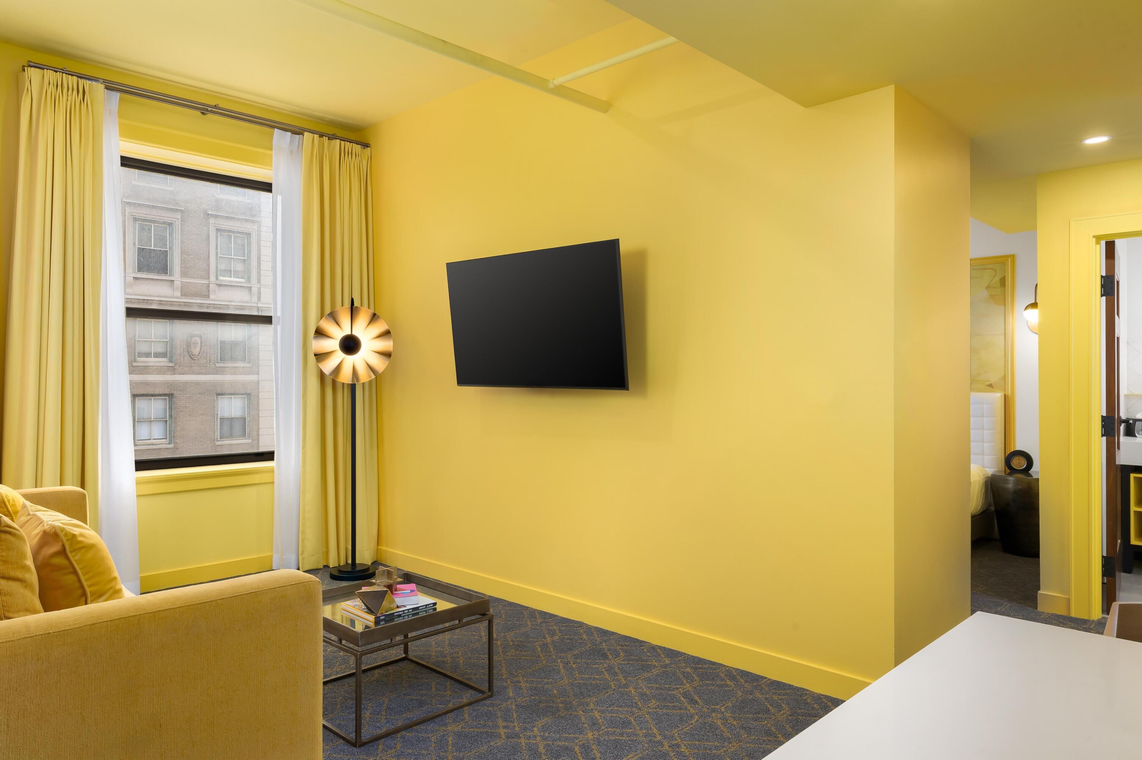 A hotel room with a yellow wall, a flat-screen TV mounted on the wall, a sofa, a small coffee table, and a window with yellow curtains.