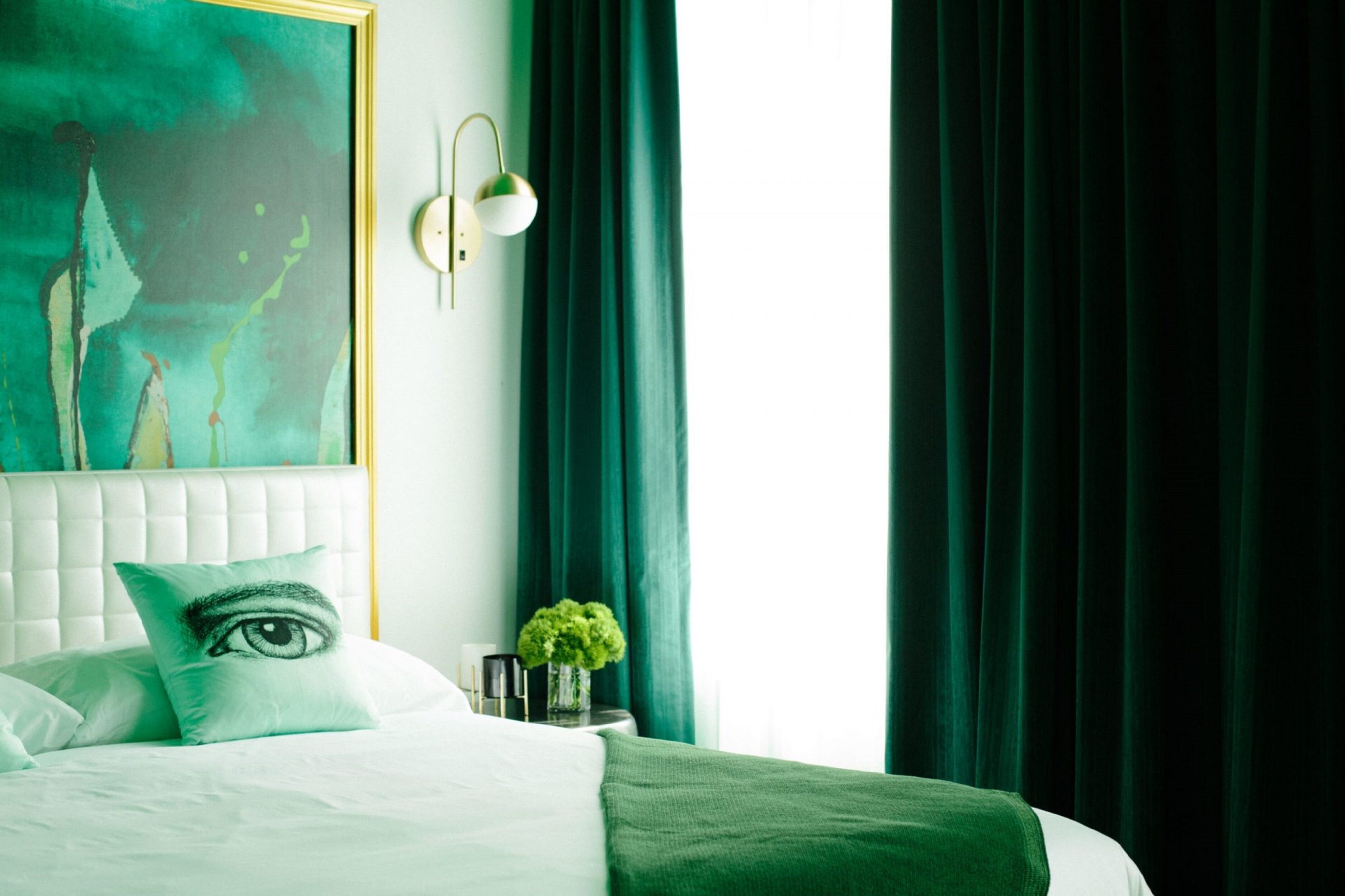 A bedroom with a bed, a wall-mounted painting, a bedside table with a lamp, and a window with curtains.