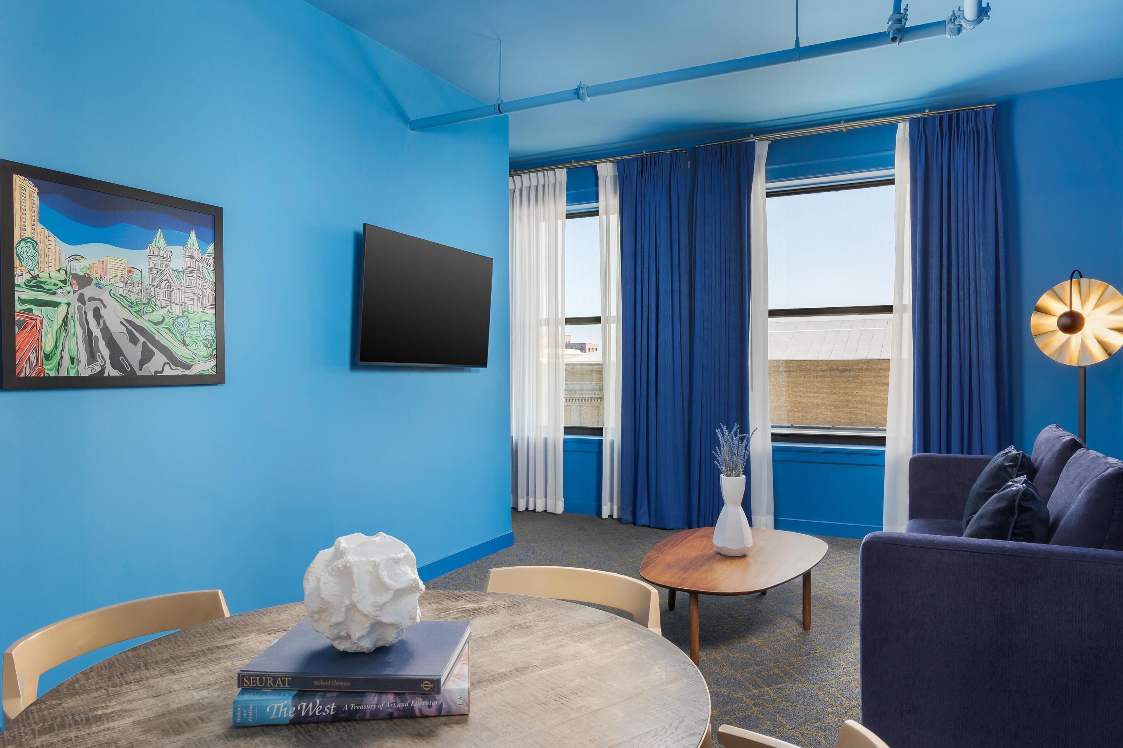 A room with a blue wall, a flat-screen TV, a painting, a sofa, a table with books, a vase with flowers, and a lamp.