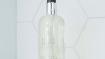 Une bouteille de savon pour les mains de la marque Molton Brown, posée sur une surface blanche, avec un mur recouvert de carreaux hexagonaux en arrière-plan.