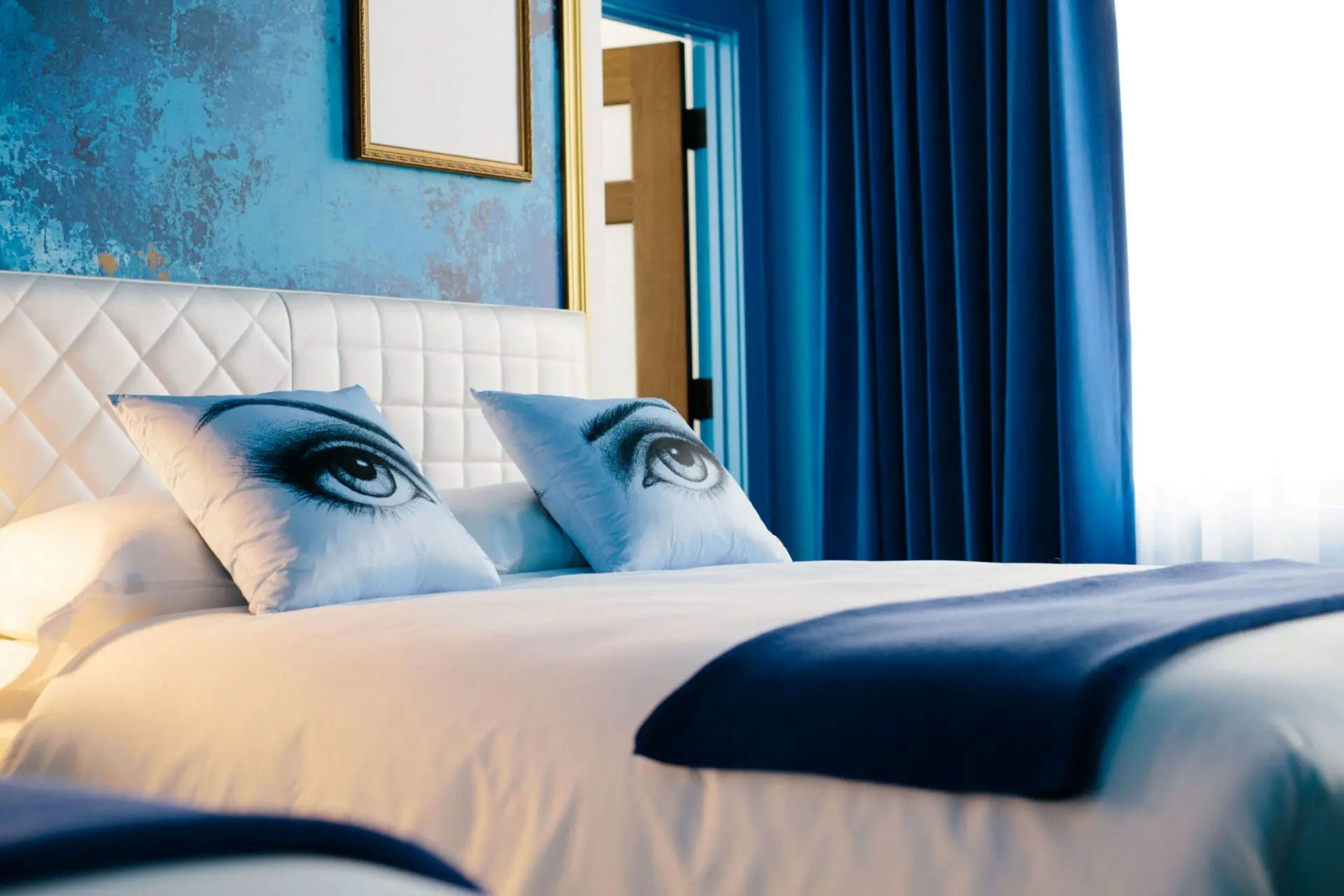 Habitación de hotel con cama blanca, dos almohadas con diseño de ojo, cortina azul y un cuadro en la pared.
