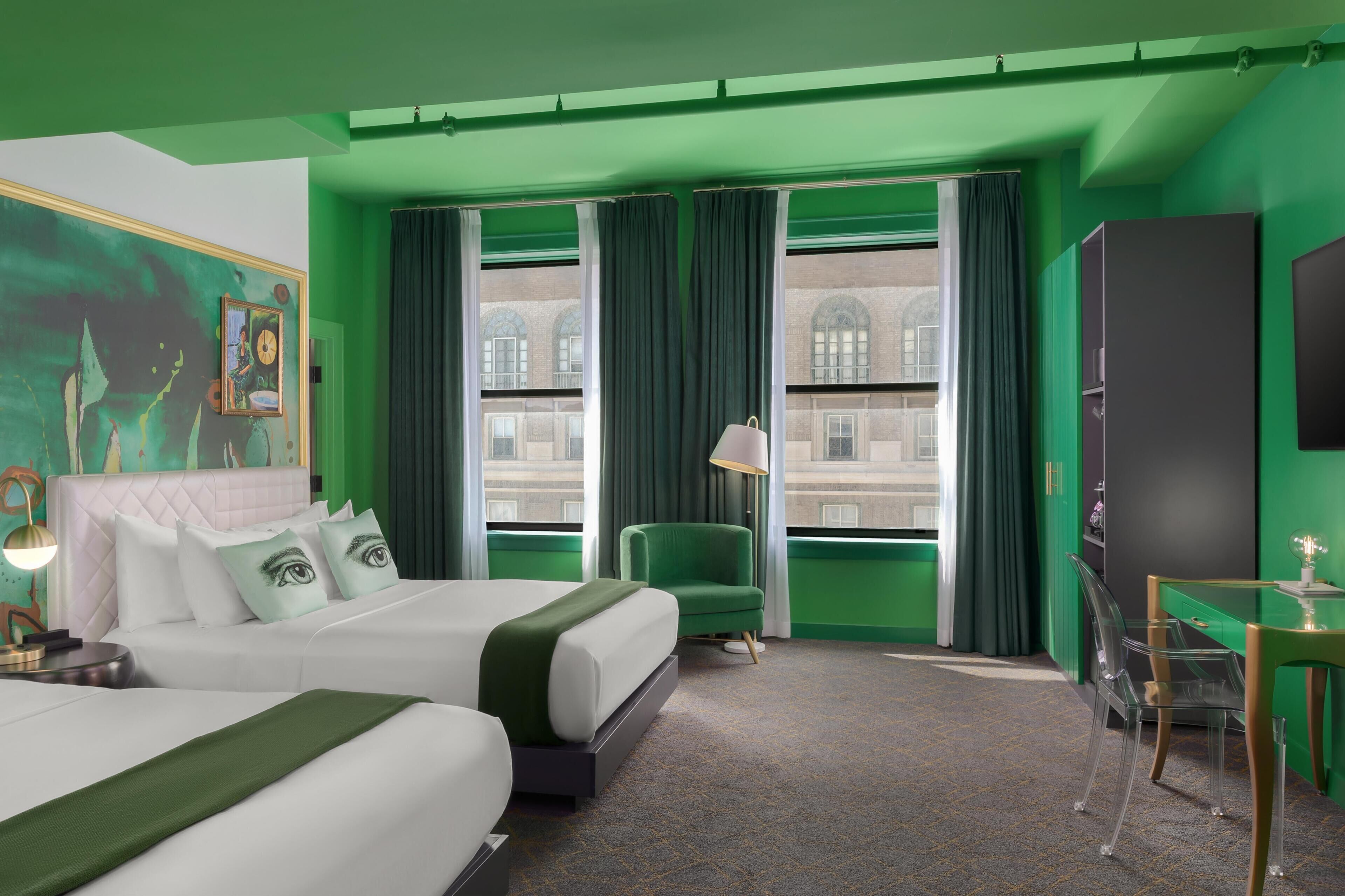 Habitación de hotel con dos camas, una silla verde, un escritorio y un ventanal con cortinas.