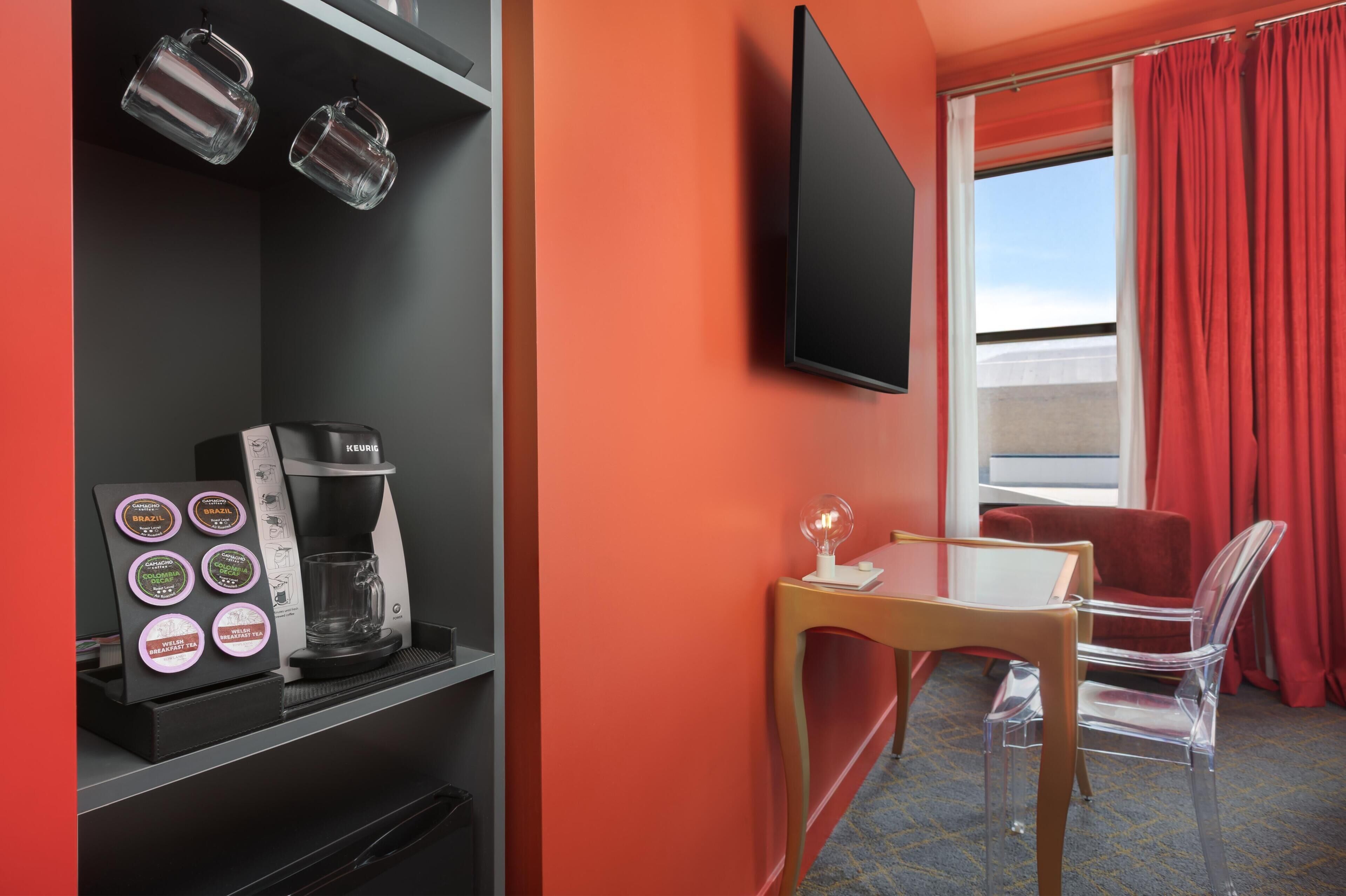 A hotel room with a coffee machine, a flat-screen TV, a table, and a chair.