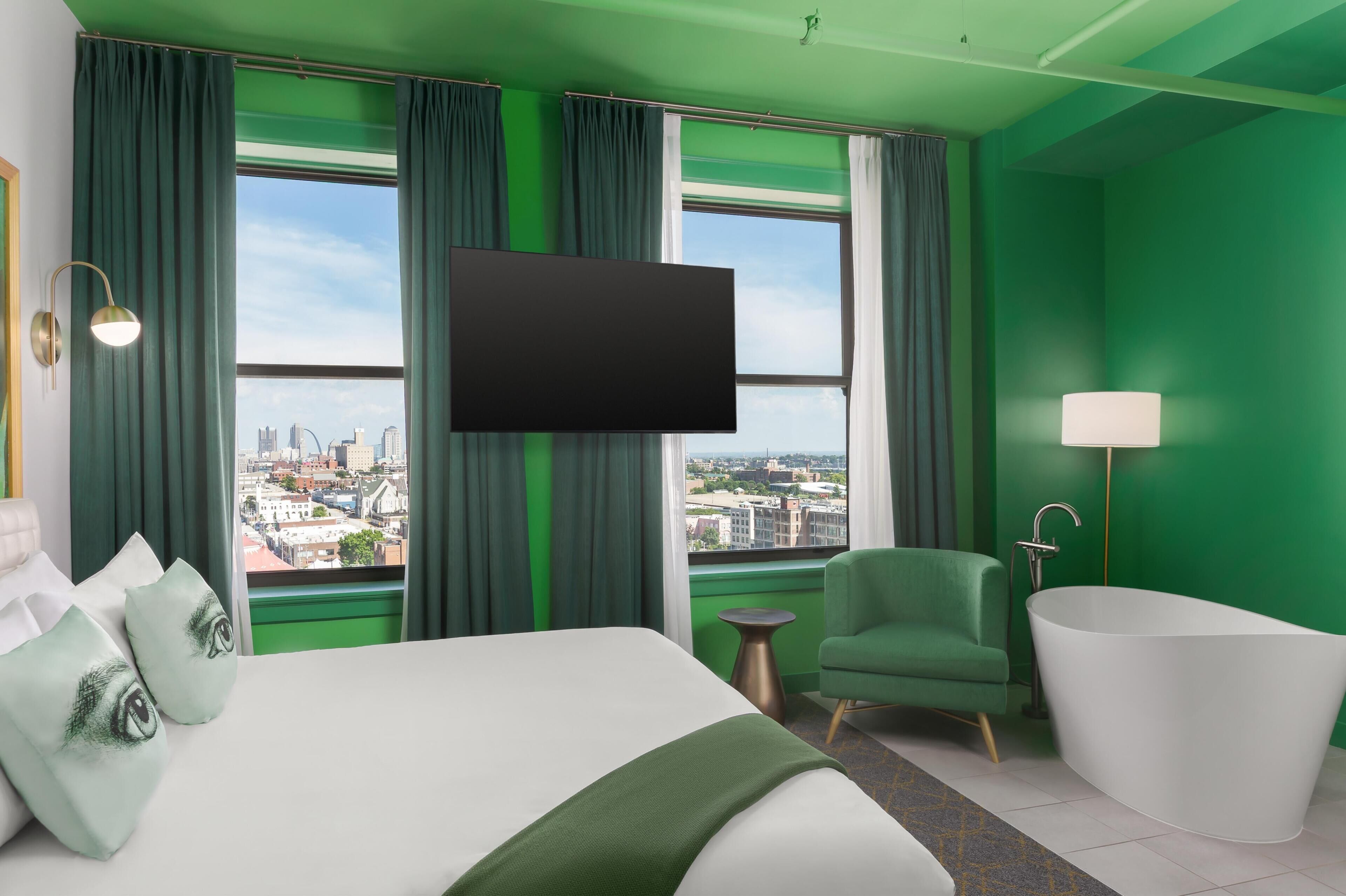 A hotel room with a large bed, a green armchair, a white bathtub, and a TV mounted on the wall.