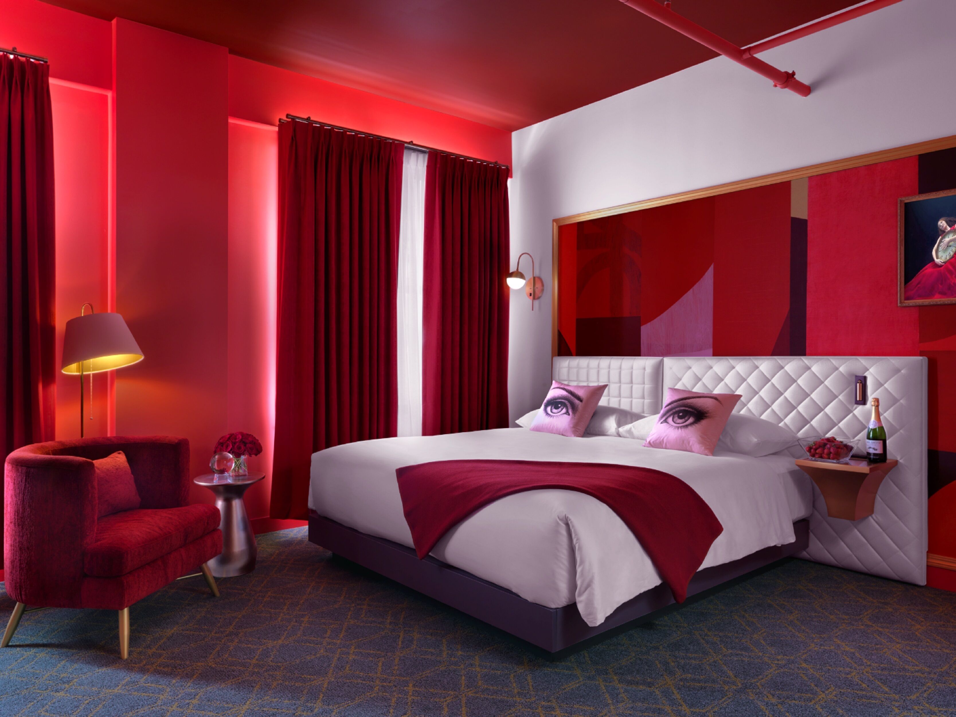 Ein modernes Hotelzimmer mit einem großen Bett, roten Vorhängen, einem roten Sessel, einem kleinen Tisch mit einer Lampe und einem Gemälde an der Wand.