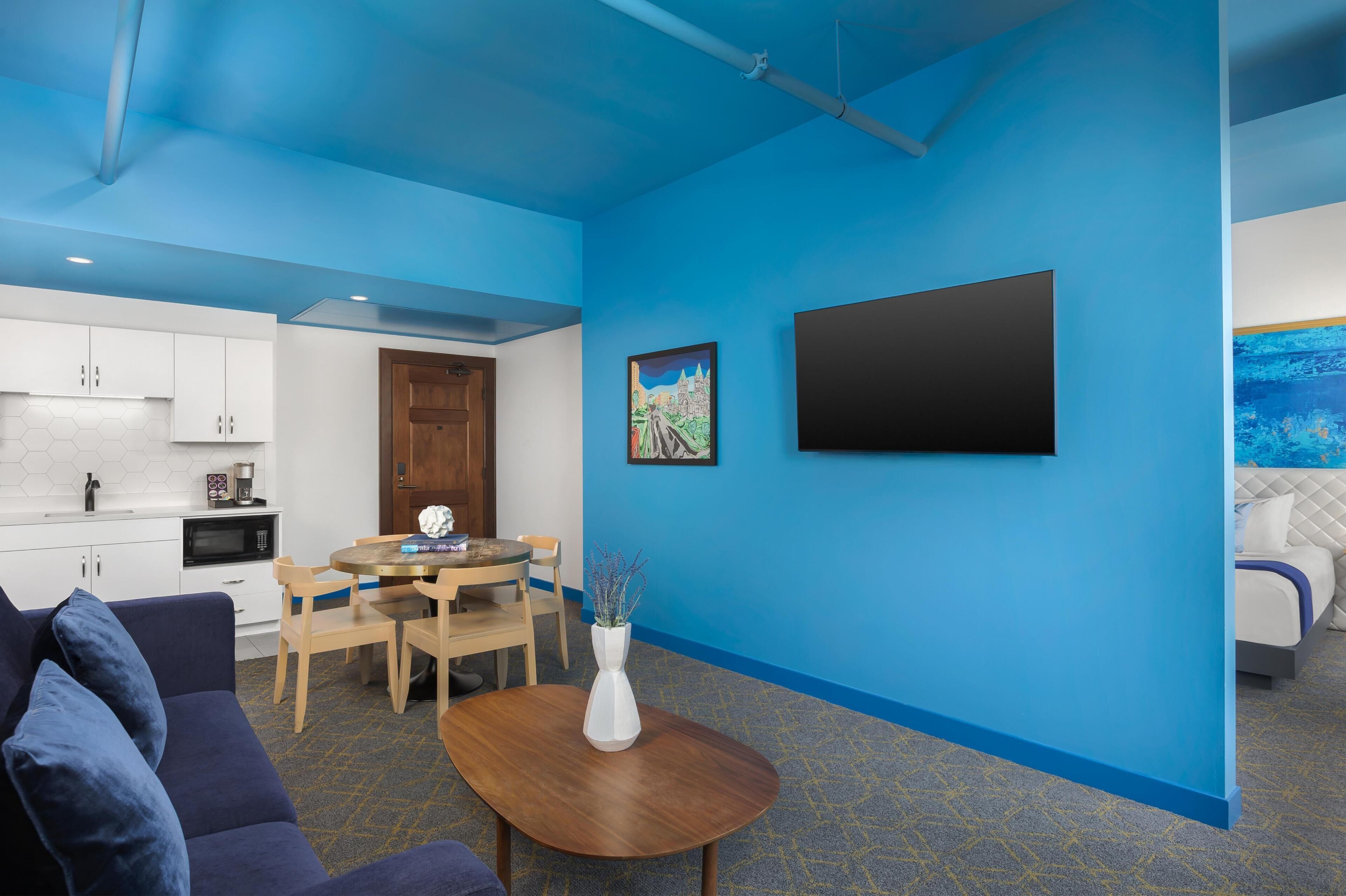 A modern hotel room with a blue accent wall, a dining area with a round table and chairs, a kitchenette, and a bed with a blue headboard.