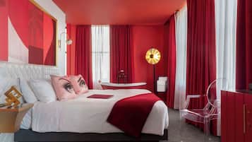 A modern hotel room with a large bed, red curtains, a round table, and a clear chair.