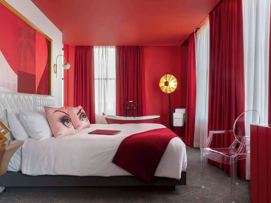 A modern hotel room with a large bed, red curtains, a round table, and a clear chair.