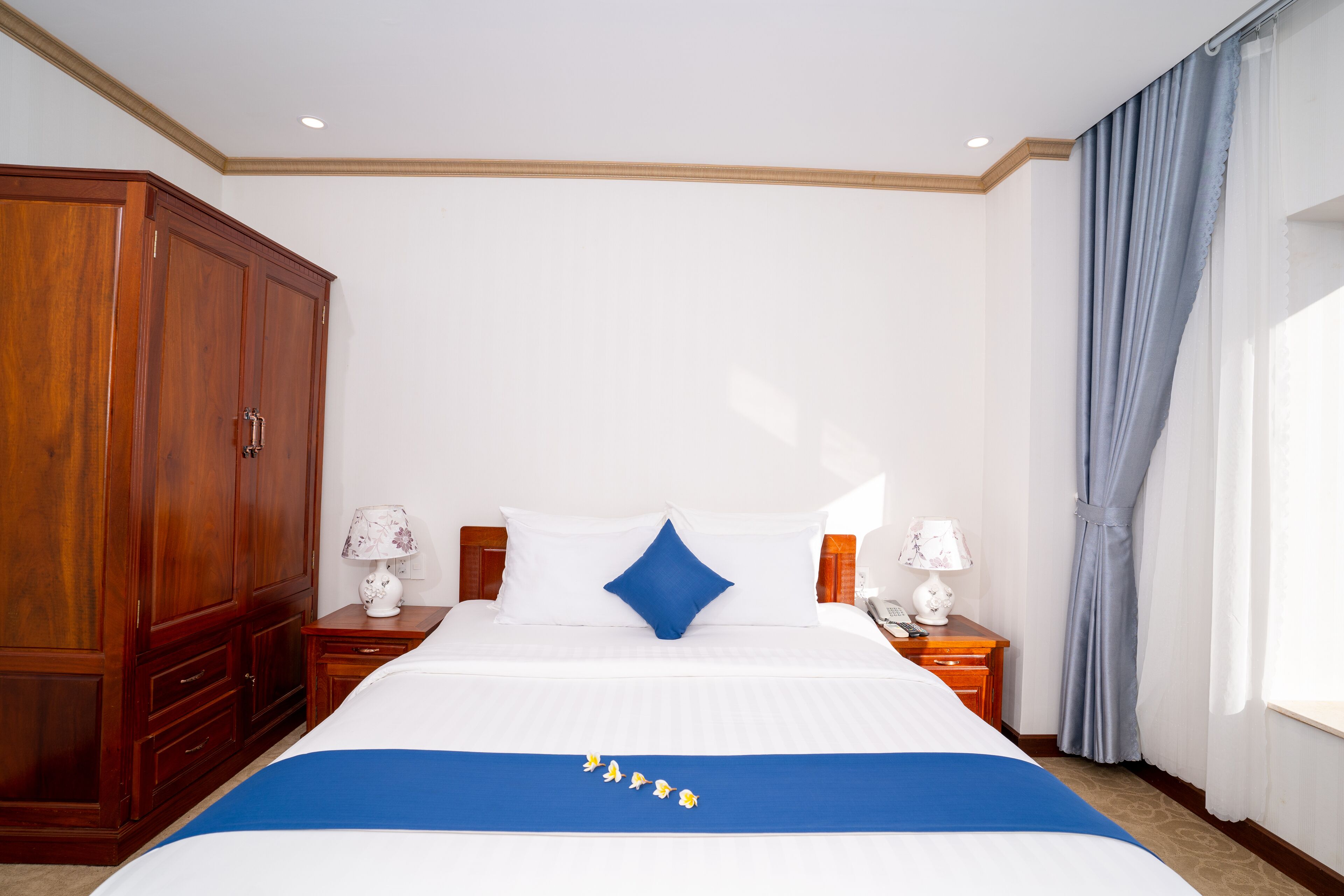 Habitación de hotel con una cama grande, un armario de madera, dos mesitas de noche con lámparas y una ventana con cortinas.