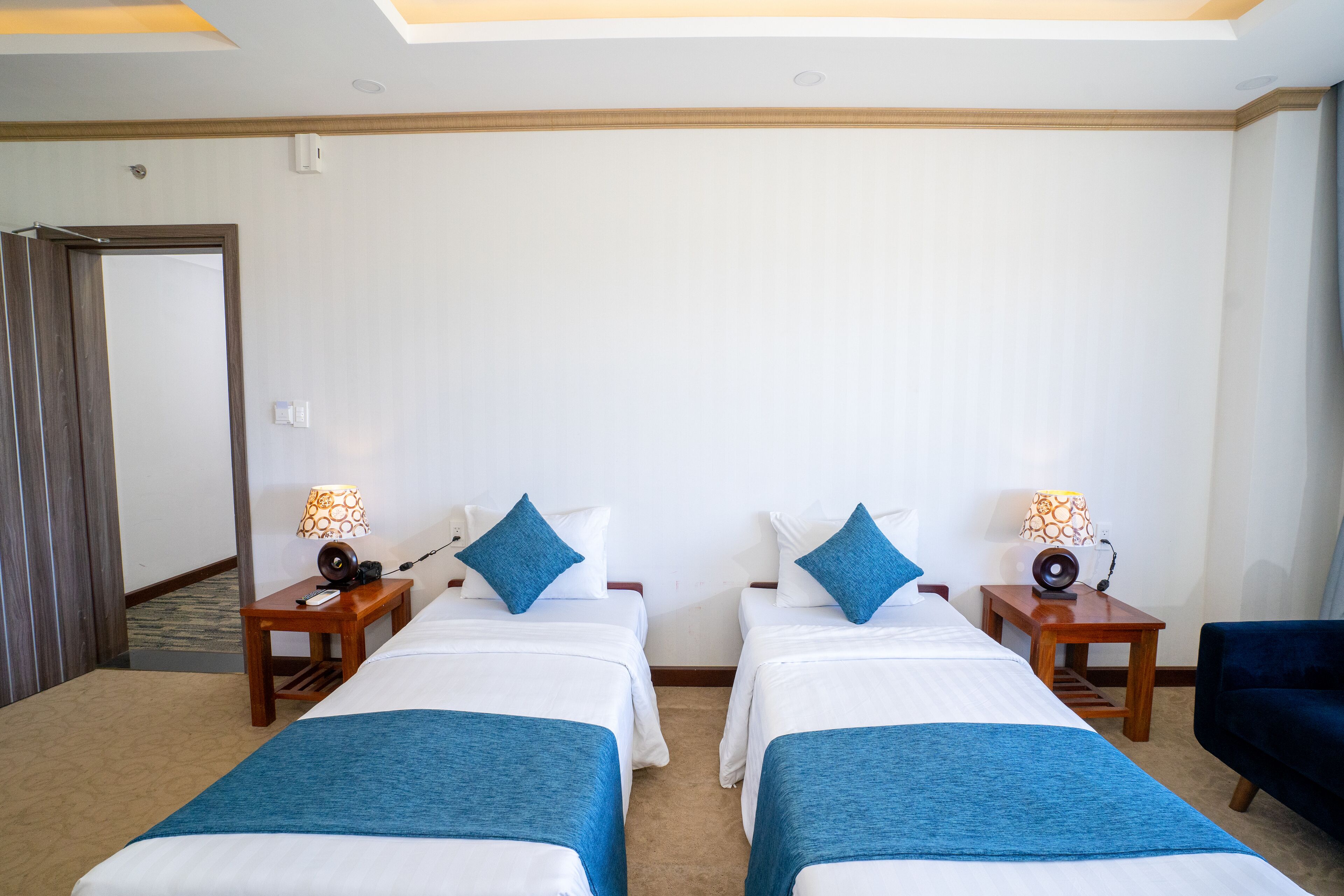 A hotel room with two beds, each with a blue throw blanket, white bedding, and matching blue pillows. There are bedside tables with lamps and a small speaker. The room has a wooden floor and a door leading to another area.