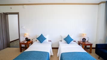 A hotel room with two beds, each with a blue throw blanket, white bedding, and matching blue pillows. There are bedside tables with lamps and a small speaker. The room has a wooden floor and a door leading to another area.
