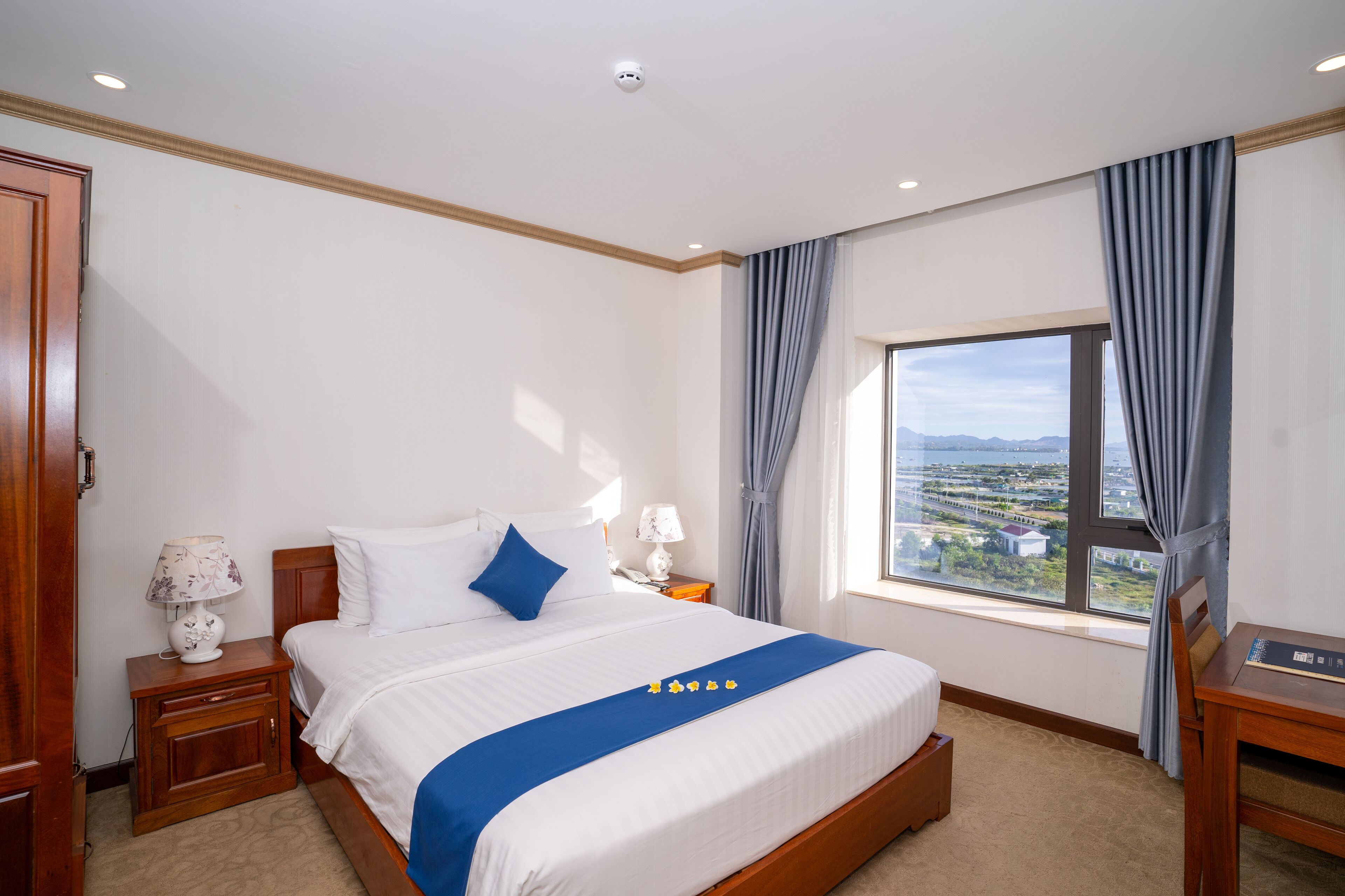 A hotel room with a large bed, a nightstand with a lamp, a wooden desk, and a view of a cityscape through a window.