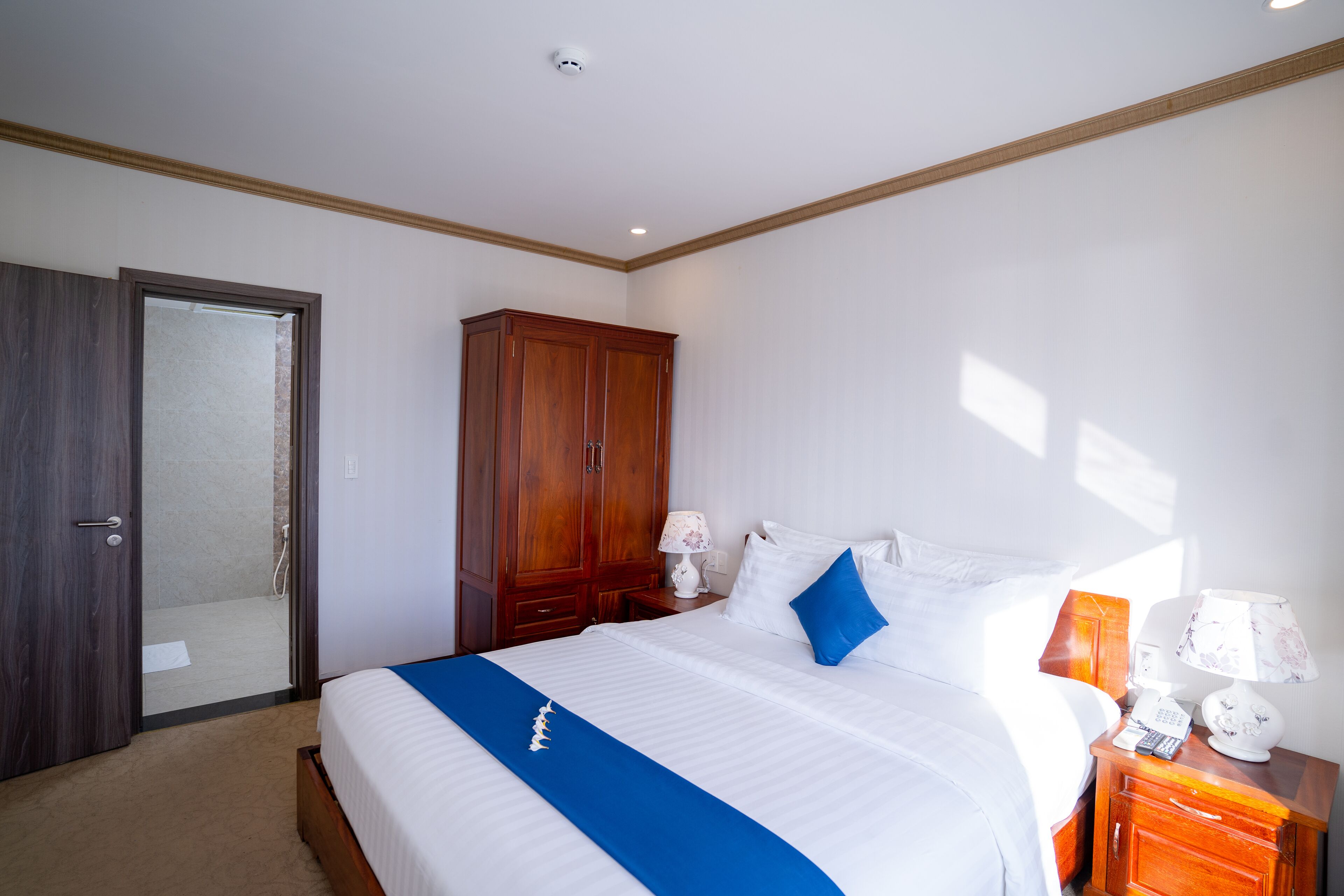 A hotel room with a bed, a wooden wardrobe, a nightstand with a lamp, and a door leading to a bathroom.
