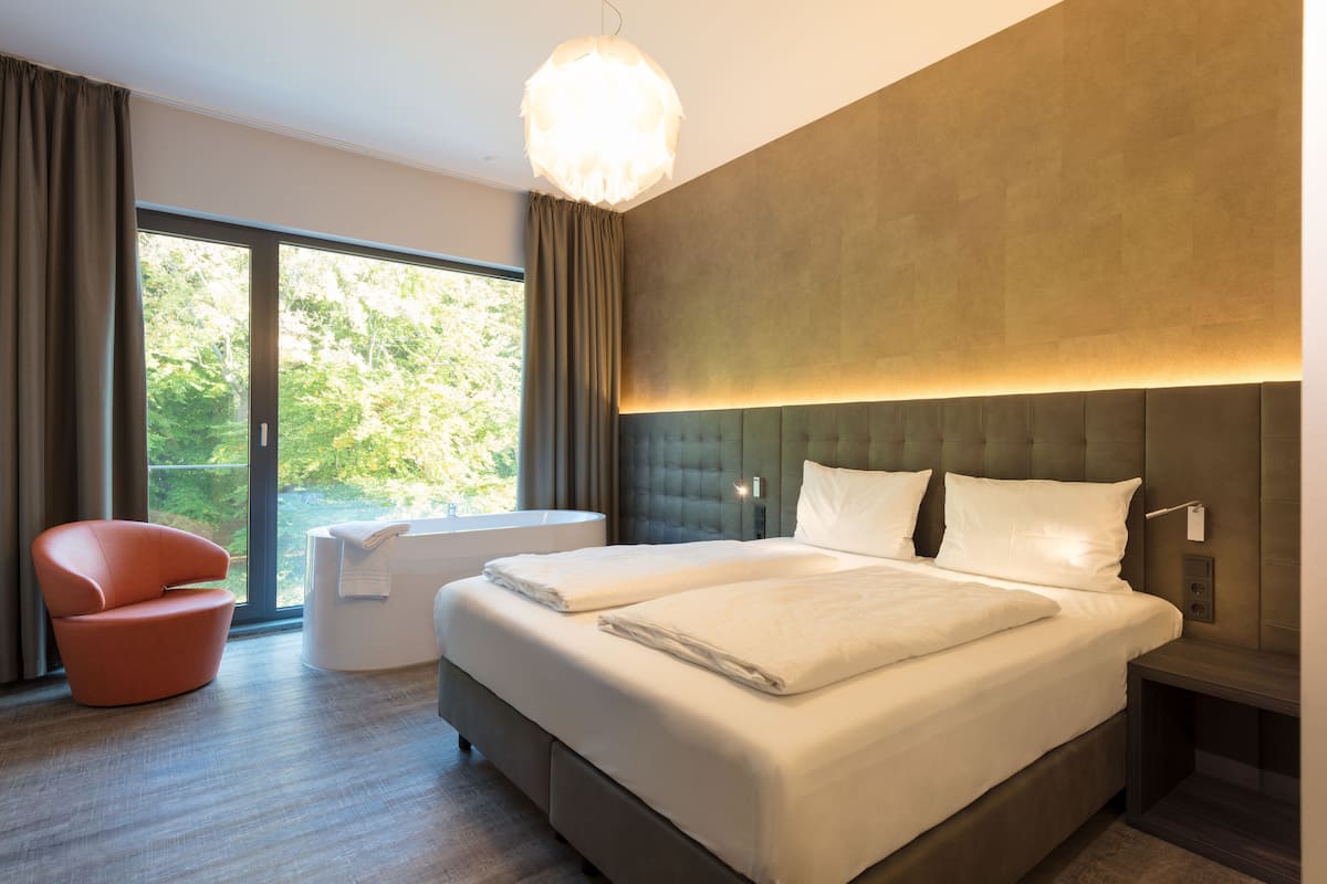 A modern hotel room with a large bed, a seating area with a chair, and a view of the outdoors.