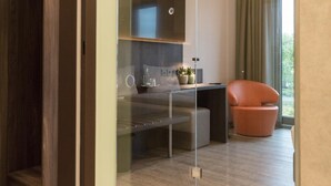 A modern, well-lit room with a glass partition, a wooden wardrobe, a seating area with a chair, and a view of greenery outside. - Deck 8 Designhotel.Soest (Soest)