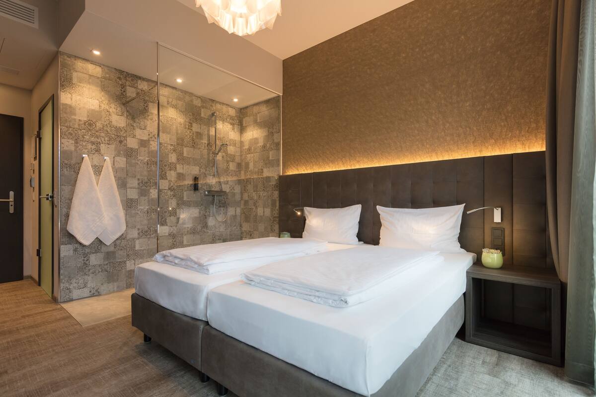 A modern hotel room with a large bed, a glass shower, and a wall-mounted light fixture.