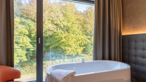 A modern bathroom with a freestanding bathtub, a view of a forest through a large window, and a cushioned seating area. - Deck 8 Designhotel.Soest (Soest)