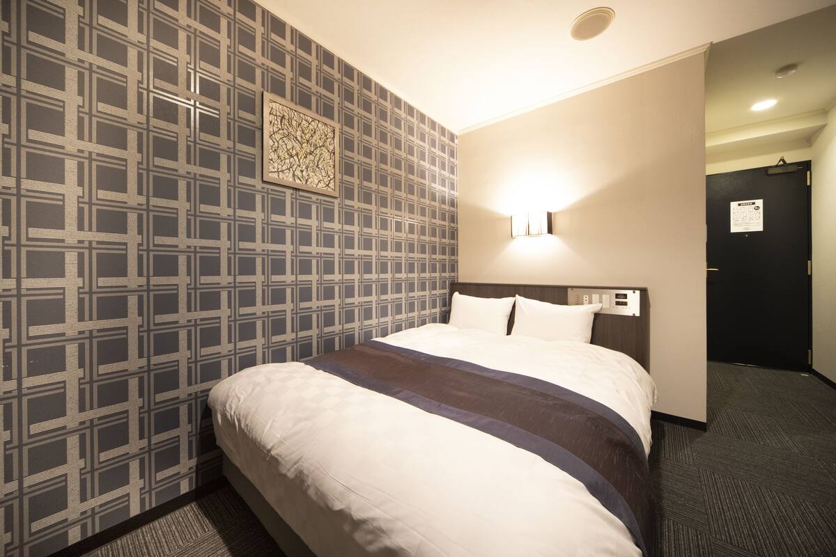 Une chambre d’hôtel avec un grand lit, un papier peint à motifs, une lampe fixée au mur et une porte avec une pancarte.