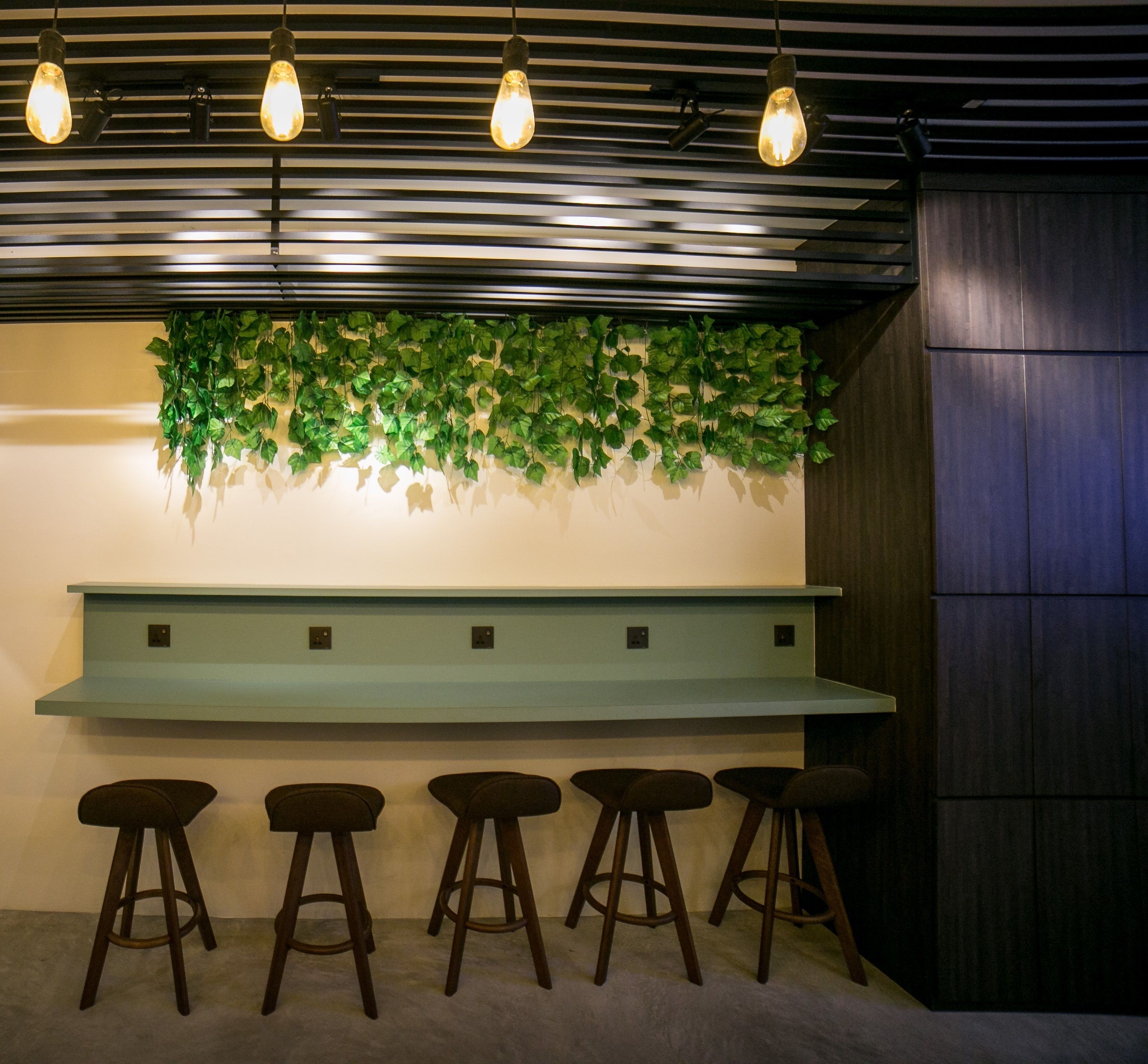 A bar area with a light blue counter, four stools, hanging light bulbs, and a green leafy wall decoration.