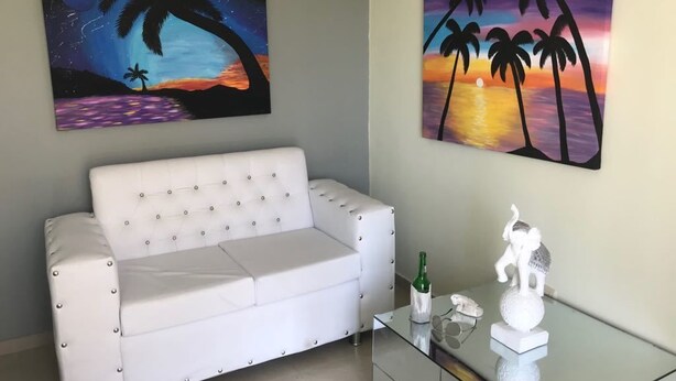 A living room with a white sofa, two paintings depicting sunsets, a glass coffee table with a bottle and a figurine. - Residencial Mia Molina (Puñal)