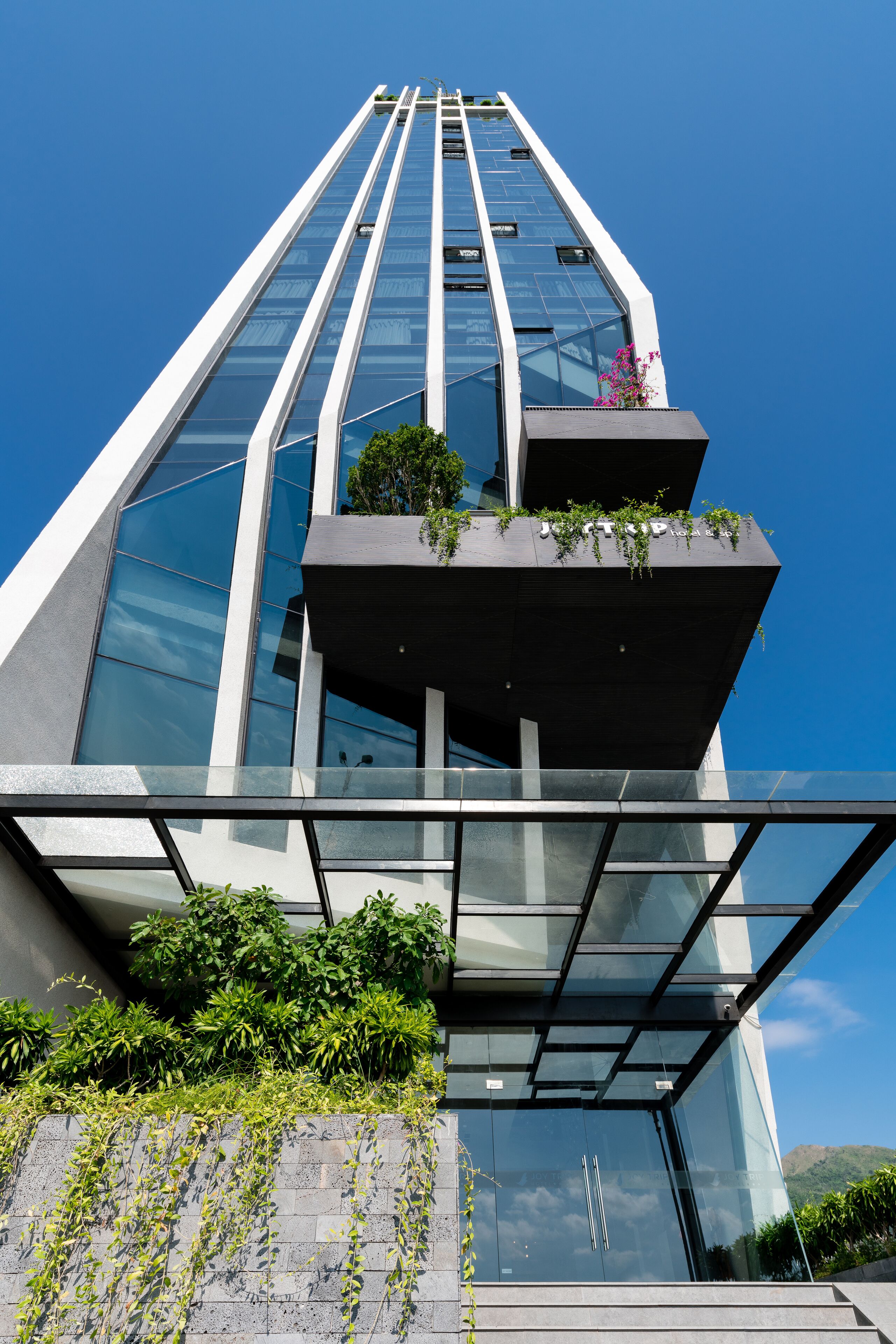 Um edifício moderno alto com fachada de vidro, varanda com plantas e céu azul claro ao fundo.