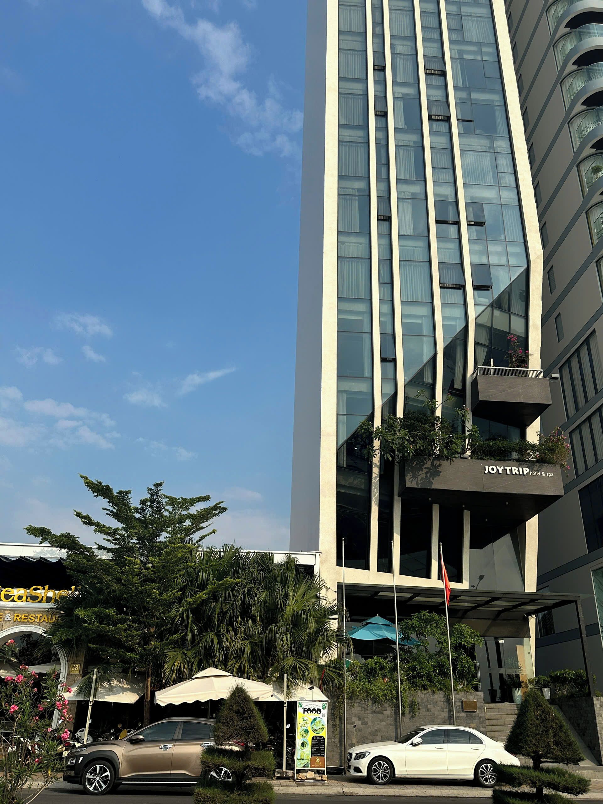 A modern hotel building with a glass facade, a visible restaurant named 'caShe' on the ground floor, and a 'JOYTRIP hotel & spa' sign on the balcony.