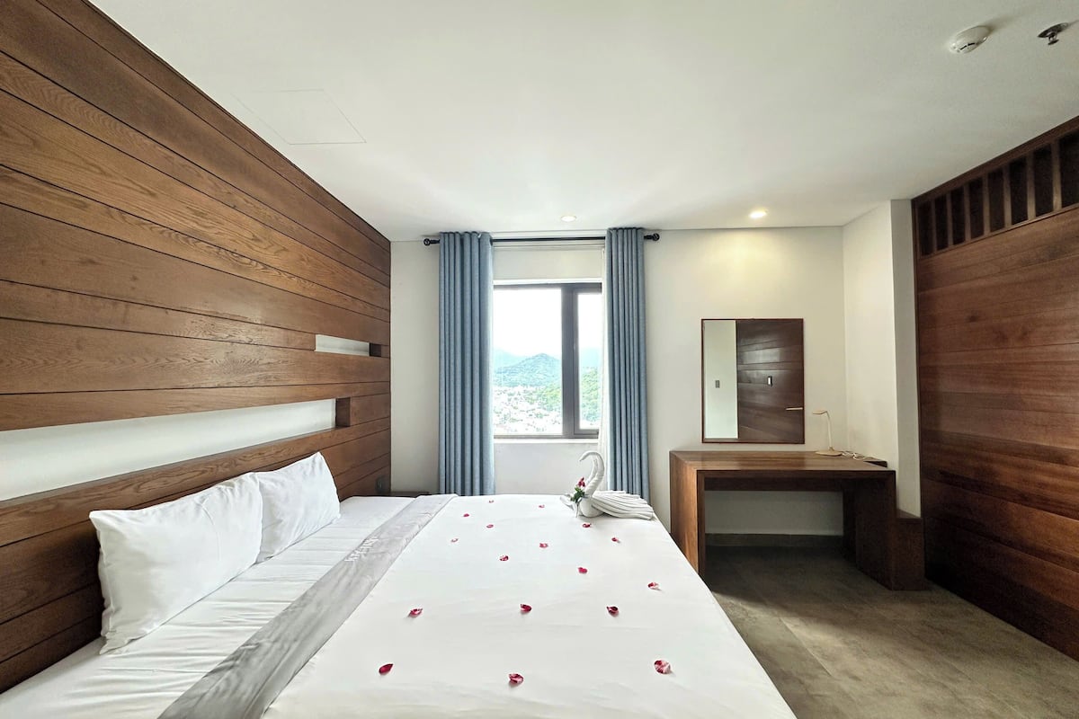 A hotel room with a wooden headboard, a bed with white linens, a bedside table, a mirror, and a view of greenery outside.