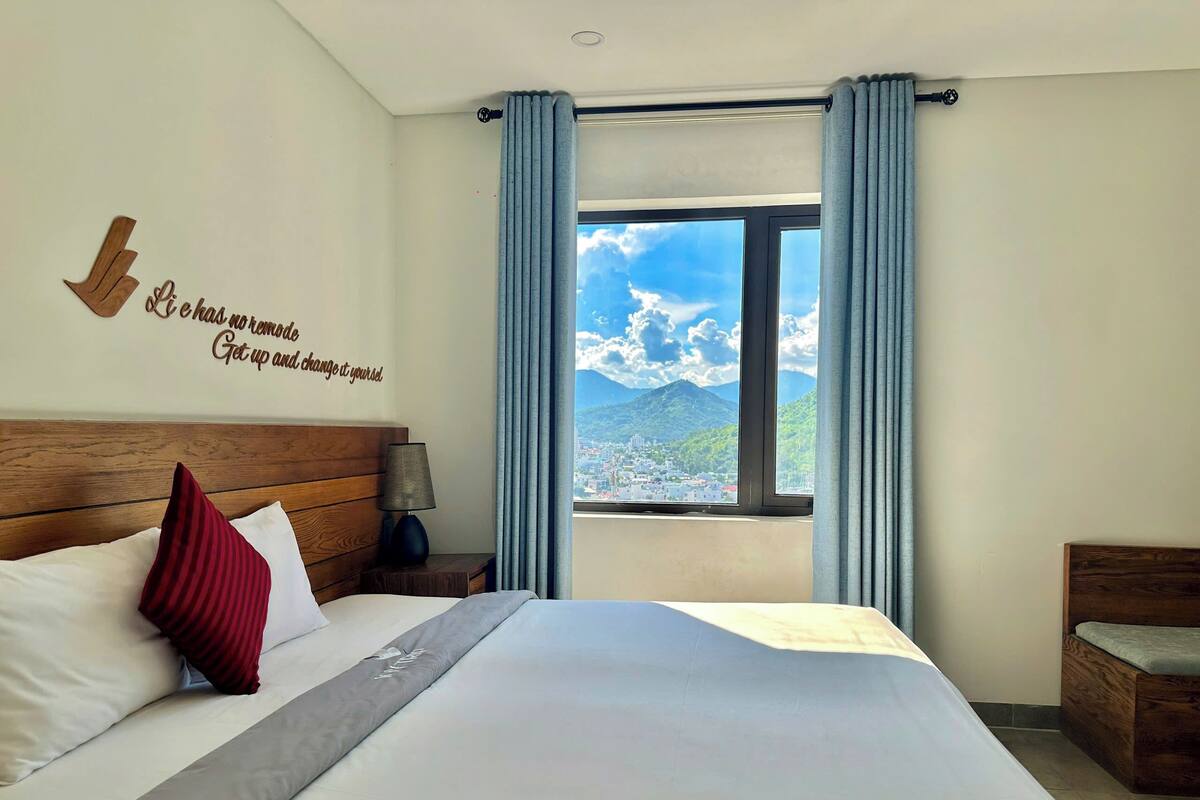 A hotel room with a bed, a nightstand, a lamp, a window with curtains, and a motivational quote on the wall.