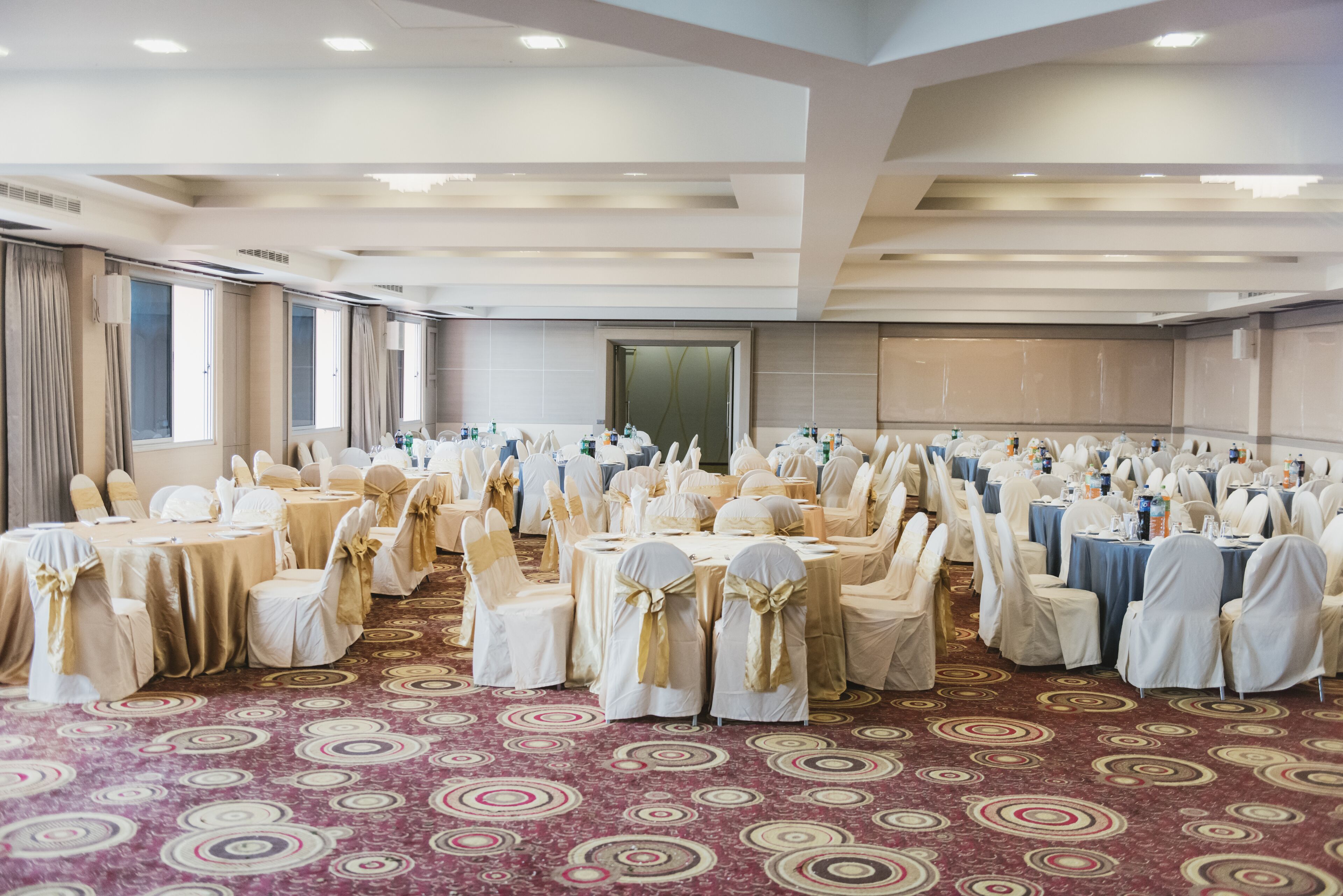 A banquet hall with round tables and chairs, a patterned carpet, and a large mirror on the wall.