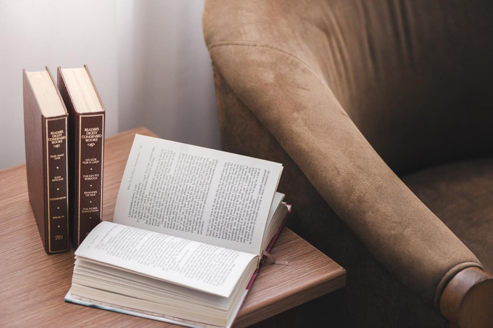 Een stapel boeken op een houten bijzettafeltje naast een bruine fauteuil.
